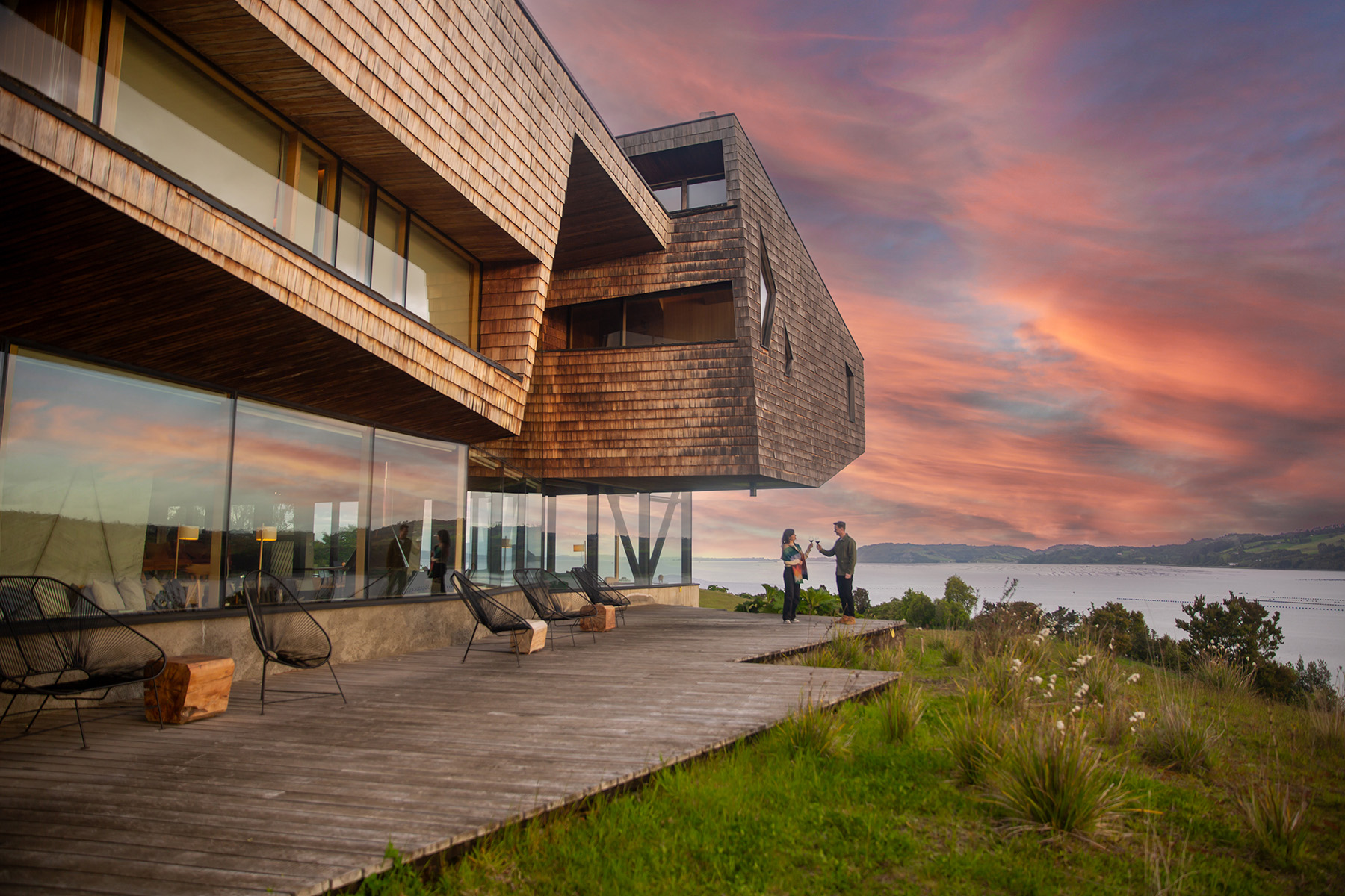 People drinking wine in front of a lodge hotel with a lake and mountains in the background