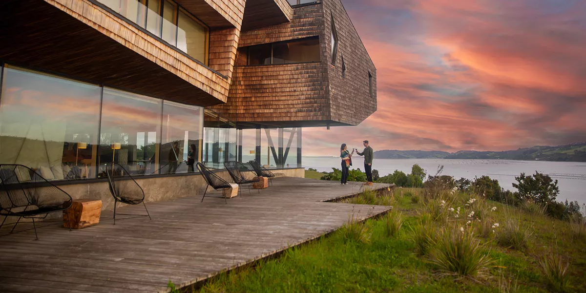People drinking wine in front of a lodge hotel with a lake and mountains in the background